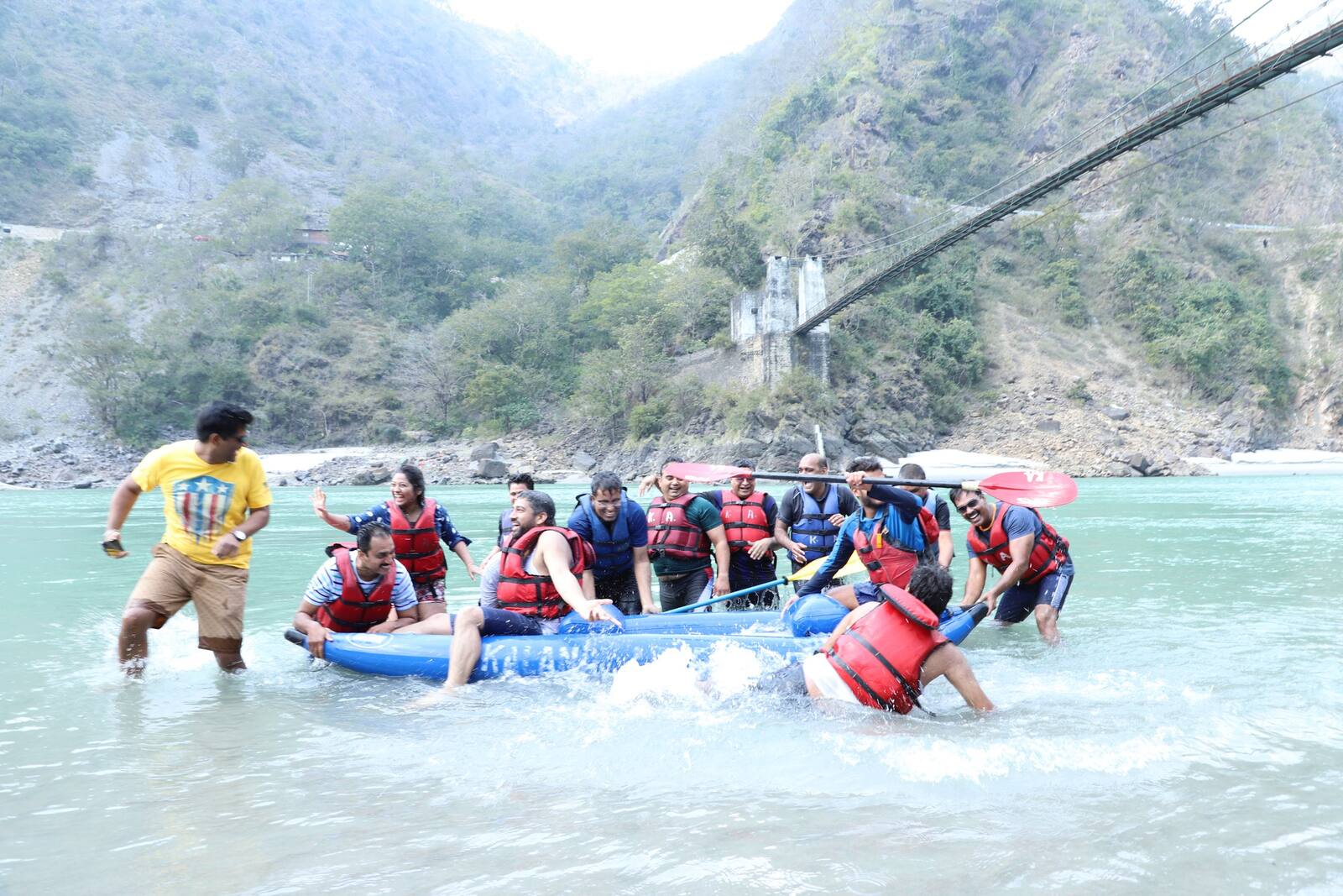 A group in life jackets pushing a raft into the Ganga, with a suspension bridge above